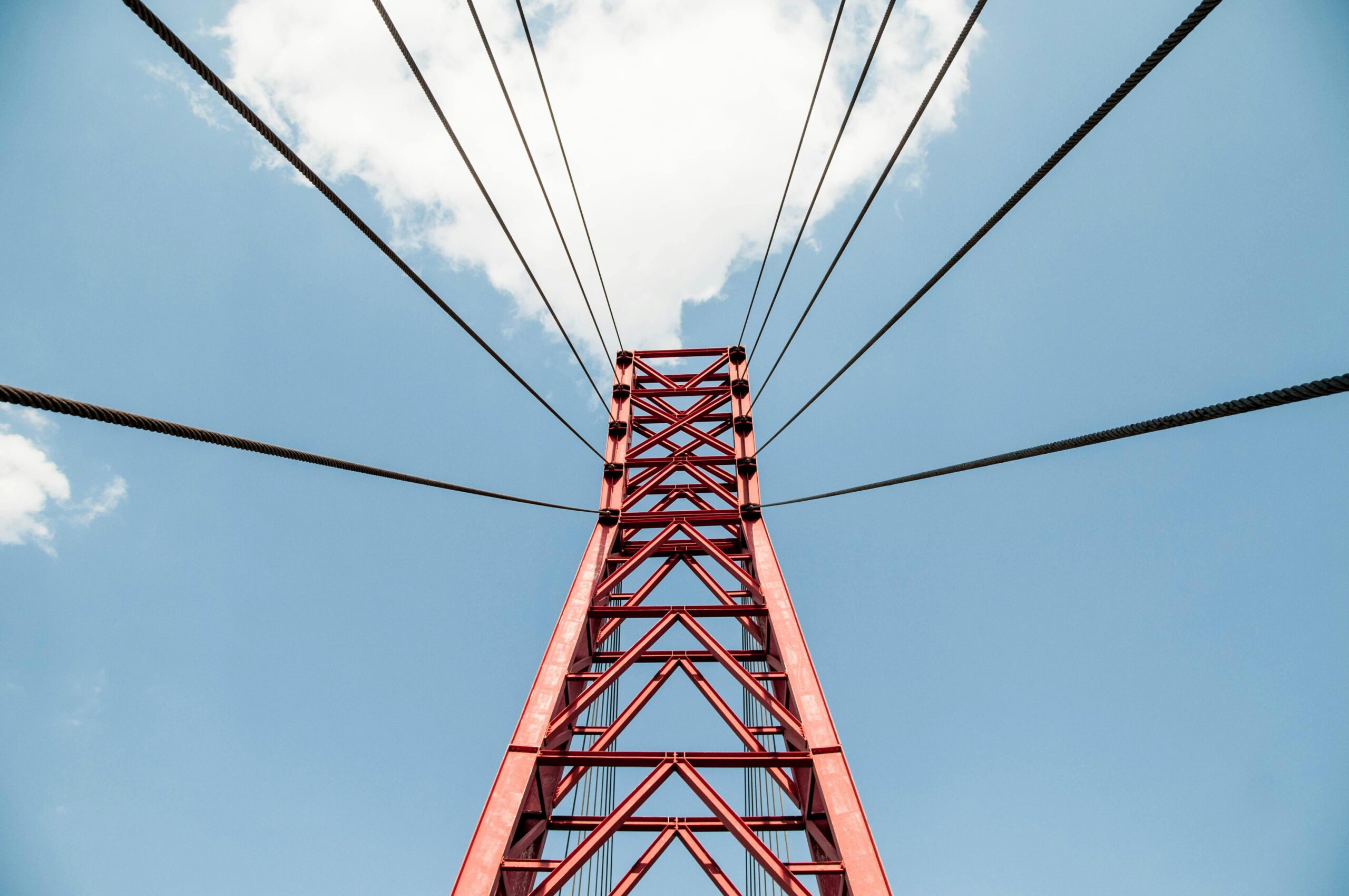 Close-up view of a red suspension bridge tower with cables, set against a bright blue sky and clouds.